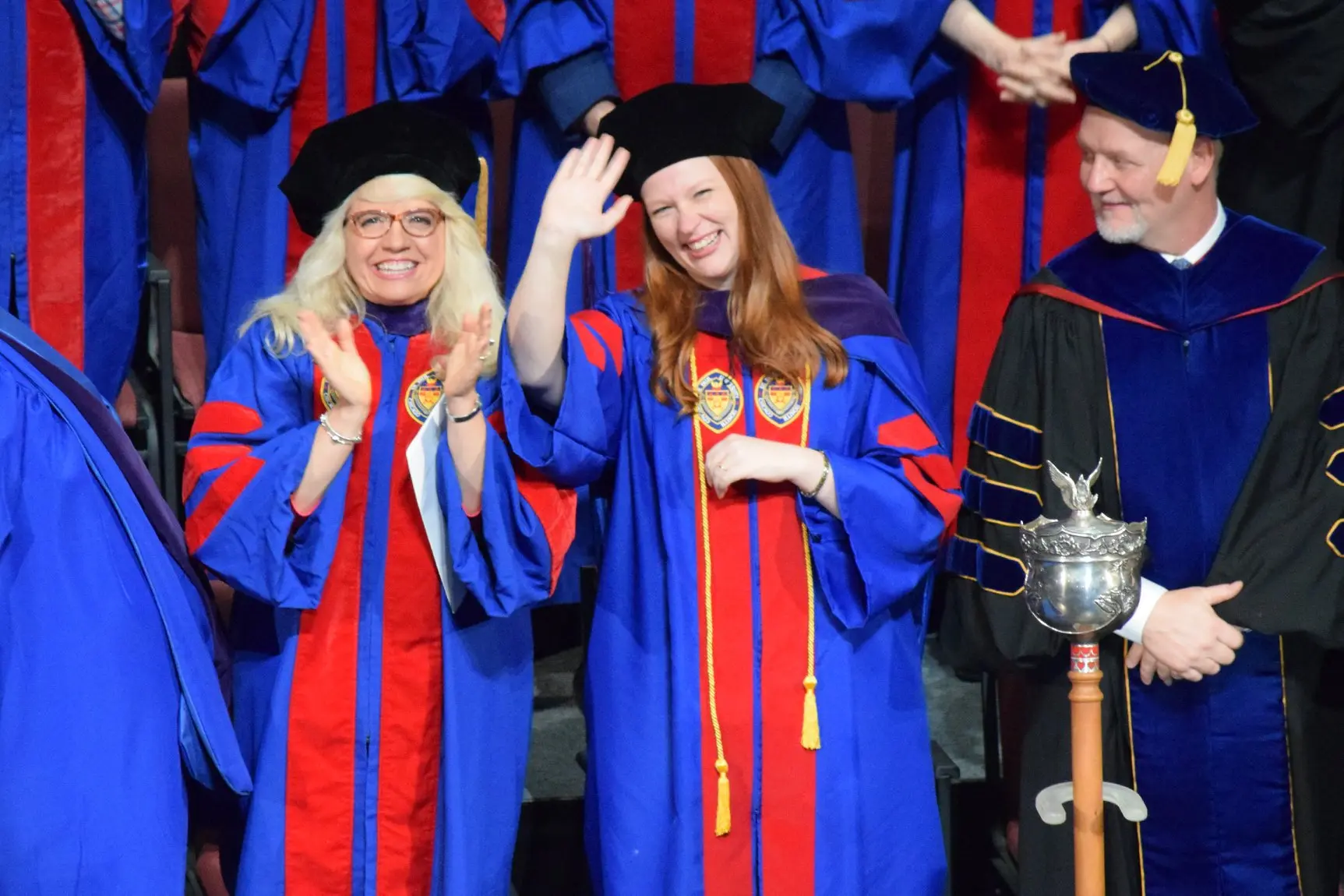 Brianna waves at the camera as she graduates from DePaul University College of Law with a Doctorate of Law Degree.  A female professor claps, while a male Dean glances at Brianna with pride.