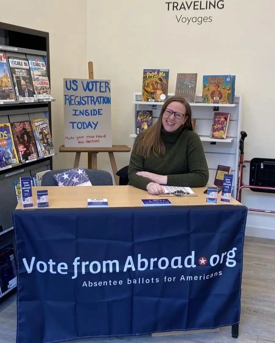 Photo of Brianna at the Institut franco-américain. She is sitting at a table that says "VotefromAbroad.org" and features an easel and pamphlets that tread "US VOTER REGISTRATION INSIDE TODAY"